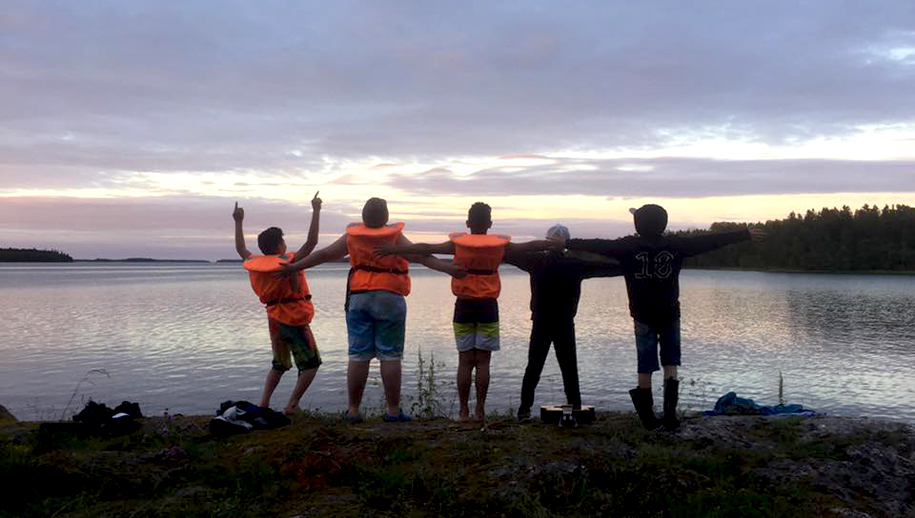 Ryggarna på fem ungdomar på en strand sträcker ut armarna mot en sjö på kvällen. 